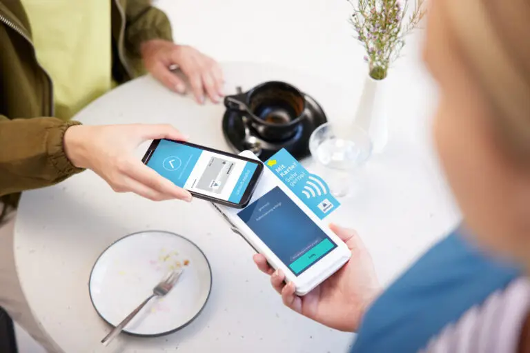 Zwei Personen nutzen ihre Smartphones für eine kontaktlose Zahlung in einem Café. Auf dem Tisch sind ein Teller mit Besteck, eine Tasse und eine Blumenvase sichtbar. Die Bildschirme der Smartphones zeigen Zahlungsanfragen und Informationen zur Transaktion.