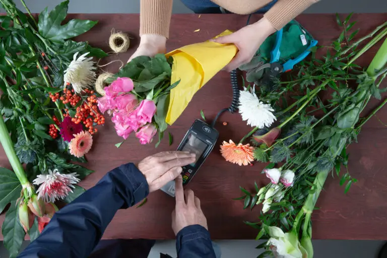 Blumenverkäuferin überreicht einen bunten Blumenstrauß in gelber Verpackung an einen Kunden, während dieser mit einem Kartenlesegerät bezahlt. Der Tisch ist mit verschiedenen frischen Blumen und Pflanzen dekoriert, darunter Rosen, Gerbera und Chrysanthemen.