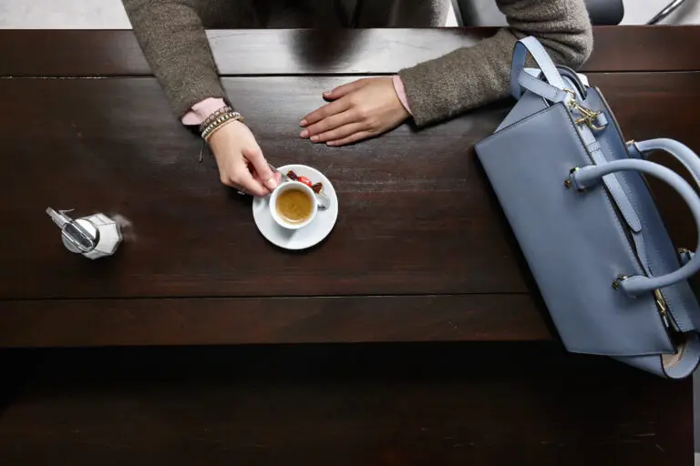 Eine Hand greift nach einer Tasse Espresso auf einem weißen Unterteller, während eine elegante Handtasche und ein Zuckergefäß auf einem dunklen Holztisch platziert sind.
