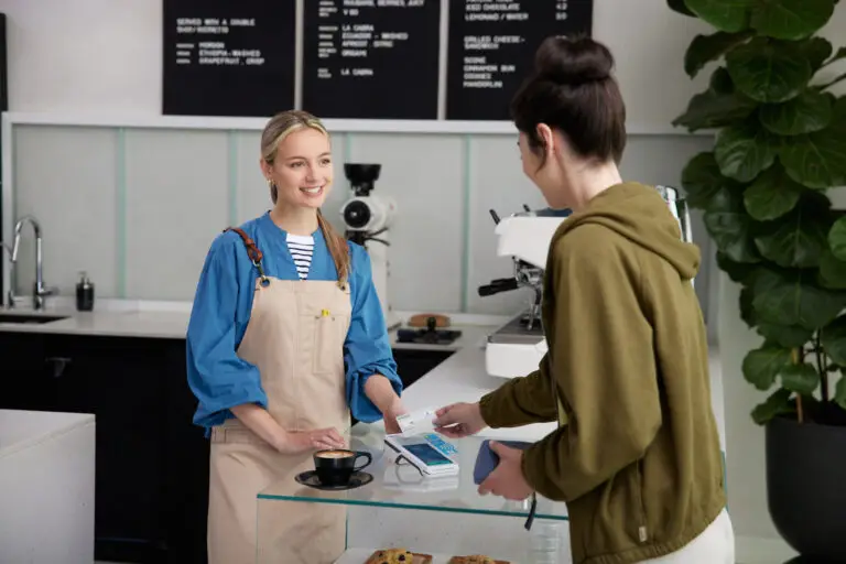 Lächelnde Barista im Café übergibt einem Kunden eine Rechnung, während dieser mit einer Karte bezahlt. Im Hintergrund sind eine Kaffeemaschine und eine Auswahl an Gebäck sichtbar.