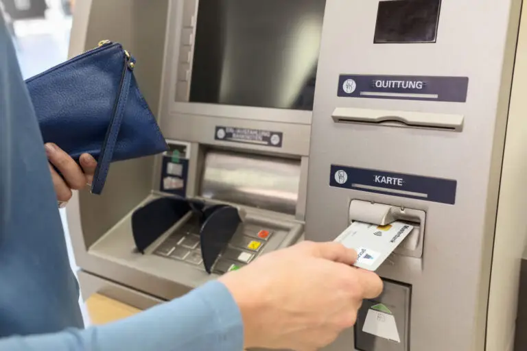Person, die an einem Geldautomaten eine Karte einführt, während sie eine blaue Geldbörse in der anderen Hand hält. Der Bildschirm des Geldautomaten ist sichtbar, und die Tasten sind gut erkennbar.