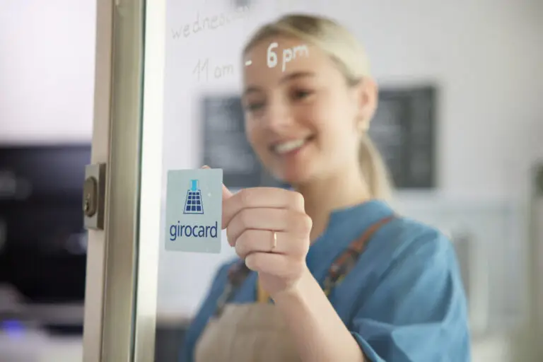 Frau klebt ein Girocard-Symbol an eine Glastür und lächelt dabei, während im Hintergrund die Öffnungszeiten sichtbar sind.