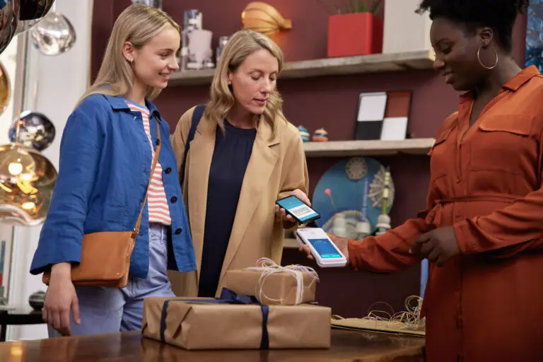 Drei Frauen in einem modernen Geschäft stehen zusammen und nutzen ihre Smartphones für eine digitale Zahlung, während Geschenke auf dem Tisch liegen. Die Atmosphäre ist freundlich und einladend, mit dekorativen Elementen im Hintergrund.