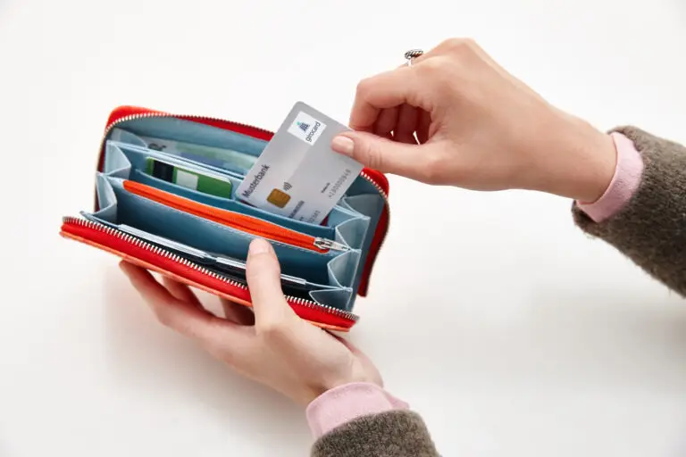 Hände halten ein rotes Portemonnaie, während eine Kreditkarte herausgenommen wird; im Inneren sind mehrere Fächer für Karten und Geldscheine sichtbar, was die praktische Nutzung und Organisation des Geldbeutels zeigt.