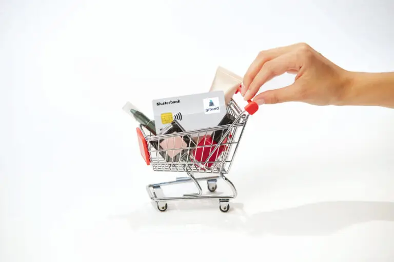 Hand, die eine Kreditkarte über einem kleinen Einkaufswagen mit Kosmetikprodukten hält. Der Einkaufswagen enthält verschiedene Schönheitsartikel wie Lippenstifte und Nagellacke, sowie eine girocard der Musterbank.