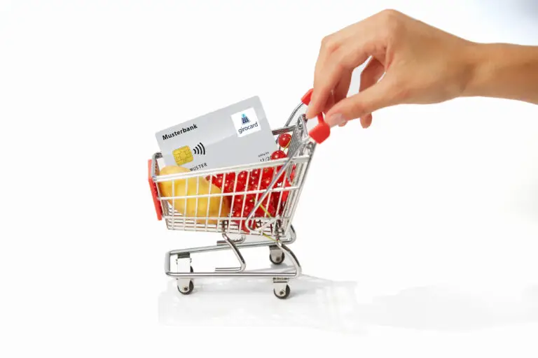 Eine Hand legt einen roten Artikel in einen Einkaufswagen, der mit frischen Lebensmitteln wie Kartoffeln und Kirschtomaten sowie einer girocard der Musterbank gefüllt ist.