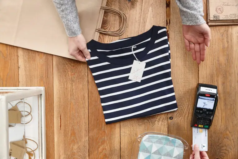 Hochwertiges gestreiftes T-Shirt in navy-blau und weiß mit Preisschild auf einem Holztisch, umgeben von einer Einkaufstasche und einem Kartenlesegerät, während der Bezahlvorgang durchgeführt wird.