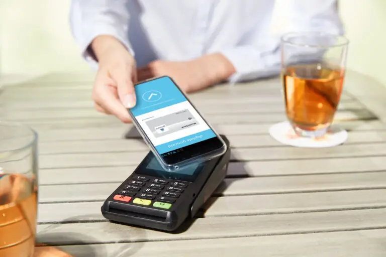 Eine Person führt eine kontaktlose Zahlung mit einem Smartphone durch, während das Gerät über ein Kartenlesegerät gehalten wird. Im Hintergrund ist ein Glas mit einem Getränk auf einem Holztisch sichtbar.