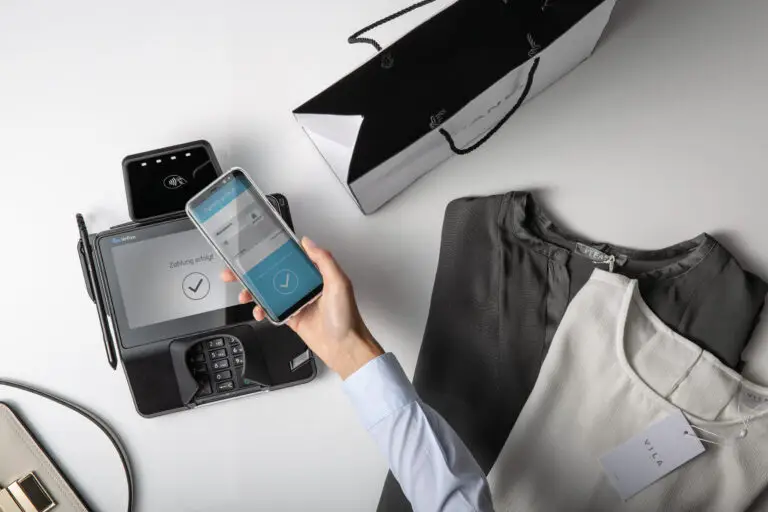 Hand mit Smartphone, das eine kontaktlose Zahlung an einem Kartenlesegerät zeigt, neben einer Einkaufstasche und zwei Oberteilen. Ideal für Online-Shopping und moderne Zahlungsmethoden.