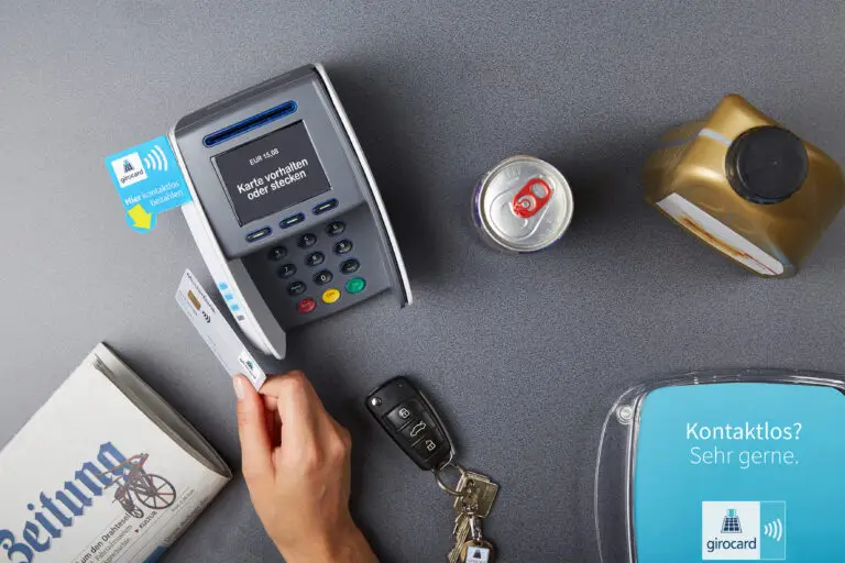 Kassenterminal mit kontaktloser Zahlungsmöglichkeit, einer Hand, die eine girocard hält, einer Getränkedose und einem Schlüsselbund auf einem Tisch.