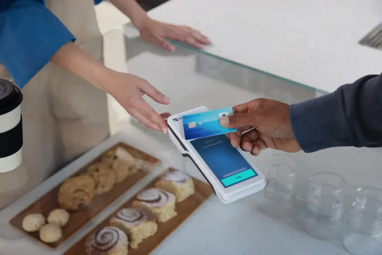 Zahlung mit Karte an einem modernen Kassensystem in einem Café, während eine Person Gebäck auf einem Holzbrett serviert.
