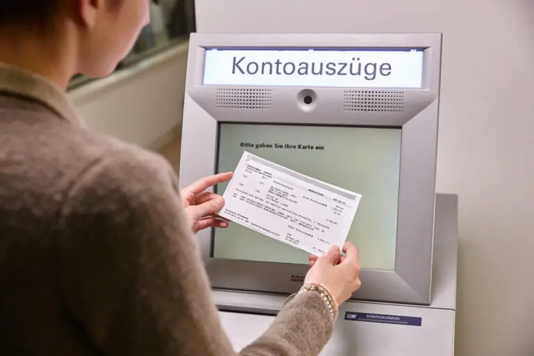 Frau benutzt einen Kontoauszugsautomaten, um einen Kontoauszug abzuholen, während sie eine Bankkarte in der Hand hält. Der Bildschirm zeigt die Aufforderung, die Karte einzugeben.