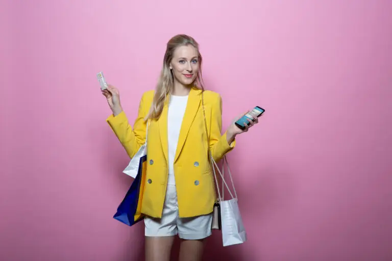 Frau mit blonden Haaren in einem gelben Blazer hält eine Kreditkarte und ein Smartphone, umgeben von Einkaufstaschen vor einem rosa Hintergrund. Ideal für Themen wie Shopping, Mode und digitale Zahlungsmethoden.
