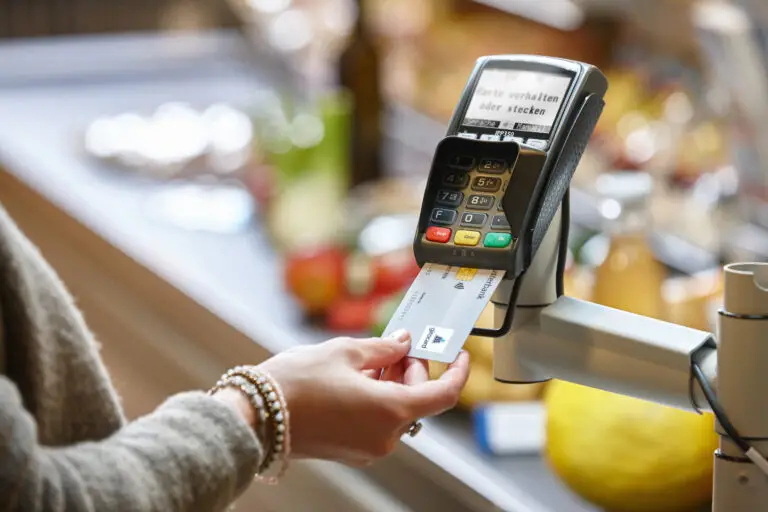 Eine Person führt eine Zahlung an einem modernen Kartenlesegerät durch, während im Hintergrund verschiedene Lebensmittel sichtbar sind. Das Display des Geräts zeigt Anweisungen zur Kartennutzung.