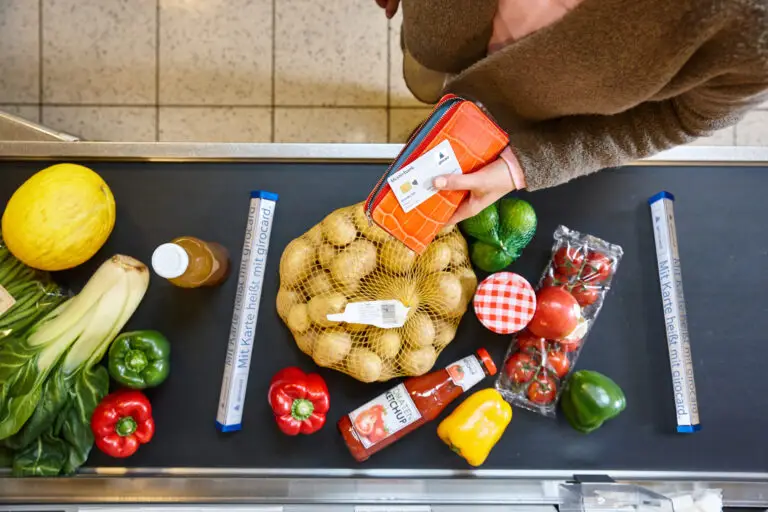 Oben auf einem Supermarktkassenband liegen frisches Gemüse und Obst, darunter Kartoffeln in einem Netz, Paprika in verschiedenen Farben, eine Flasche Saft, Ketchup und eine Hand mit einer bunten Geldbörse.