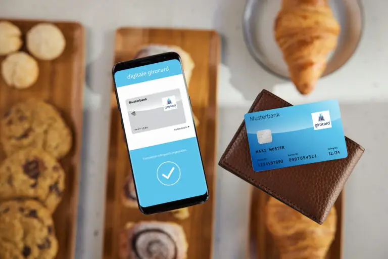 Digitale Girocard auf einem Smartphone über einem Tisch mit Keksen und einem Croissant, zeigt die Benutzeroberfläche für eine Transaktion der Musterbank.