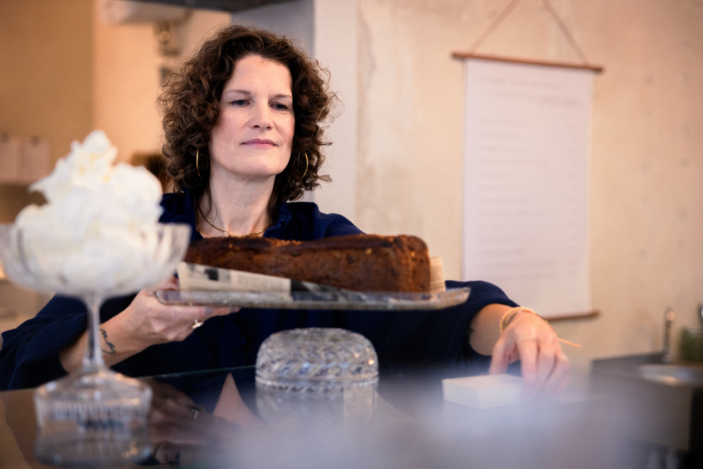 Frau mit lockigem Haar präsentiert einen frisch gebackenen Kuchen auf einem Glasteller in einem Café, während im Vordergrund eine Schüssel mit Sahne zu sehen ist.