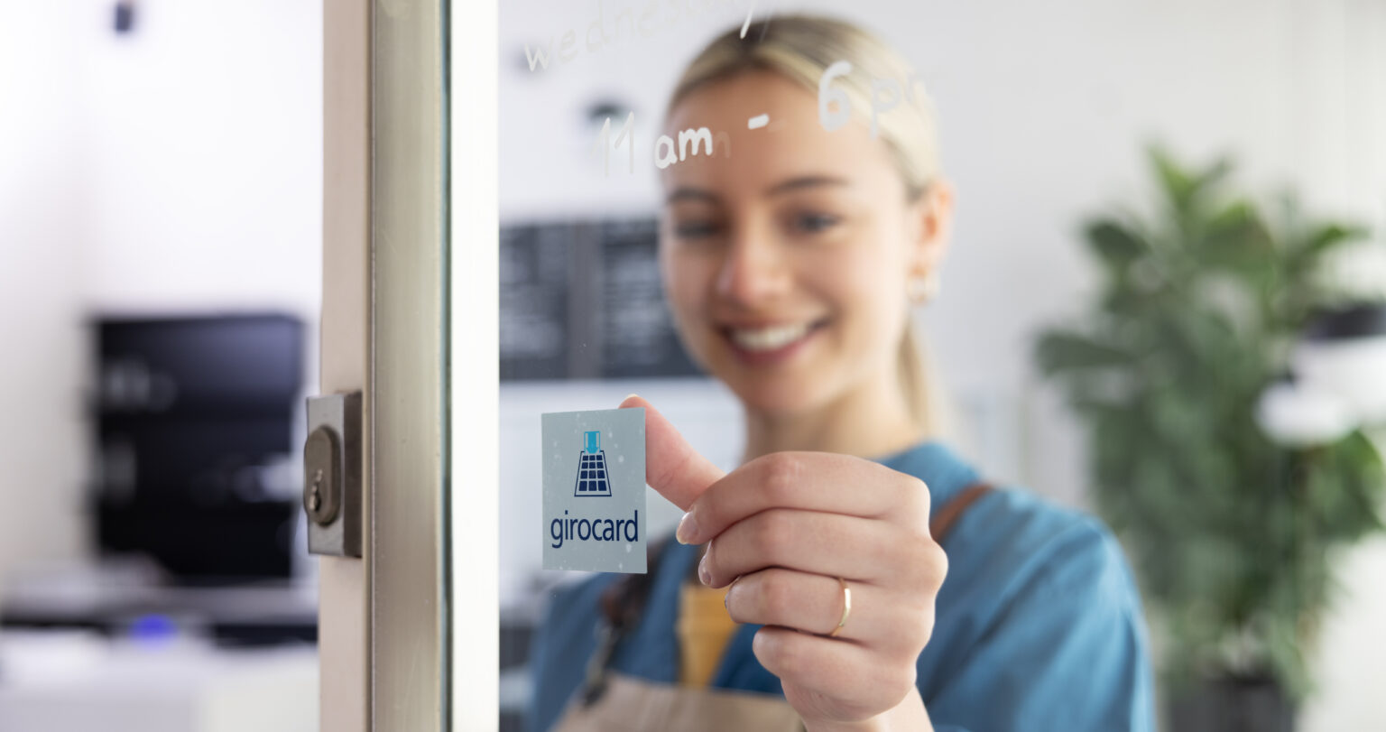 Eine lächelnde Frau bringt ein Girocard-Symbol an die Glastür eines Geschäfts an, während im Hintergrund Informationen zu den Öffnungszeiten sichtbar sind.
