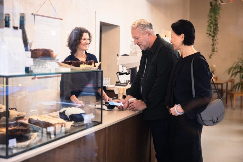 Girocard-Point-of-Sale-Szene in einer Café-Bäckerei: zwei Kunden zahlen am Tresen, eine Barista bedient den Kassentresen hinter einer Glasvitrine mit Kuchen und Kaffeemaschine im Hintergrund.
