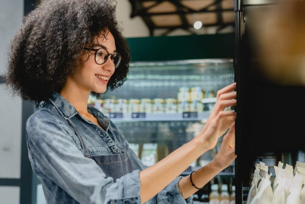 Junge Frau mit lockigem Haar und Brille, die in einem modernen Geschäft Regale mit Produkten durchsucht, lächelnd und interessiert.