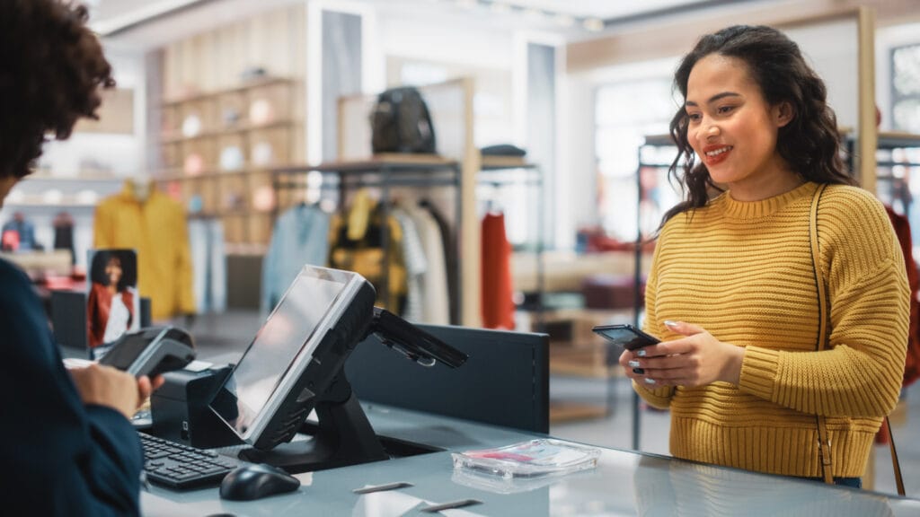 Frau in einem Modegeschäft an der Kasse, die lächelnd mit einem Smartphone in der Hand steht, während die Kassiererin einen Zahlungsvorgang durchführt. Im Hintergrund sind verschiedene Kleidungsstücke und Accessoires sichtbar.