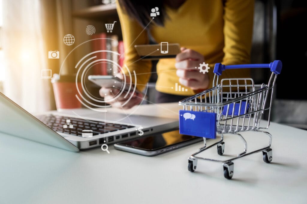 Frau nutzt Smartphone und Laptop für Online-Shopping, während ein kleiner Einkaufswagen und digitale Symbole für E-Commerce und Kommunikation im Vordergrund sichtbar sind.