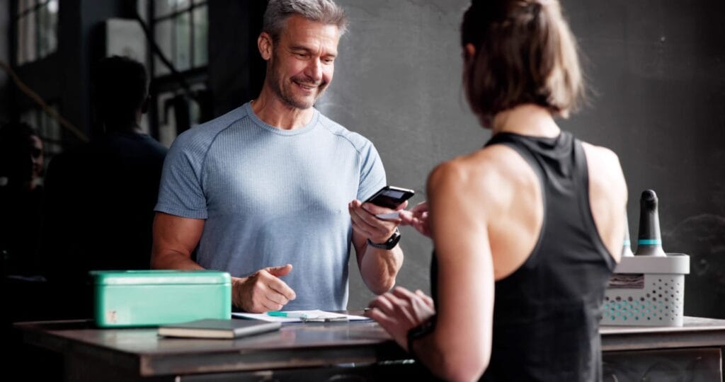 Ein Mann mit blauem T-Shirt zeigt einer Kundin an einem Tisch ein Smartphone, während ein Notizblock und eine grüne Box sichtbar sind.