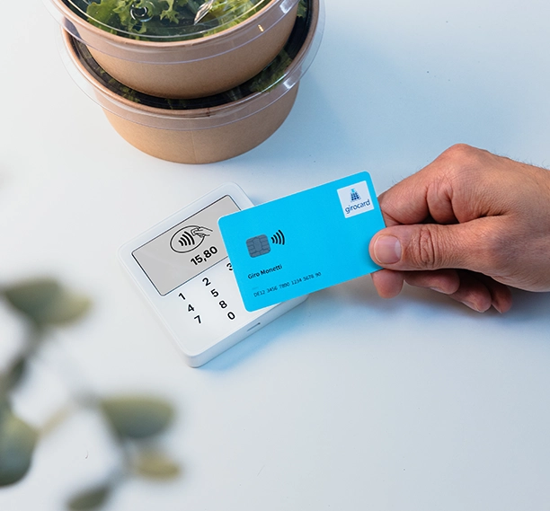 Hand hält eine girocard an ein kleines Kartenterminal, das den Betrag 15,80 Euro anzeigt.
