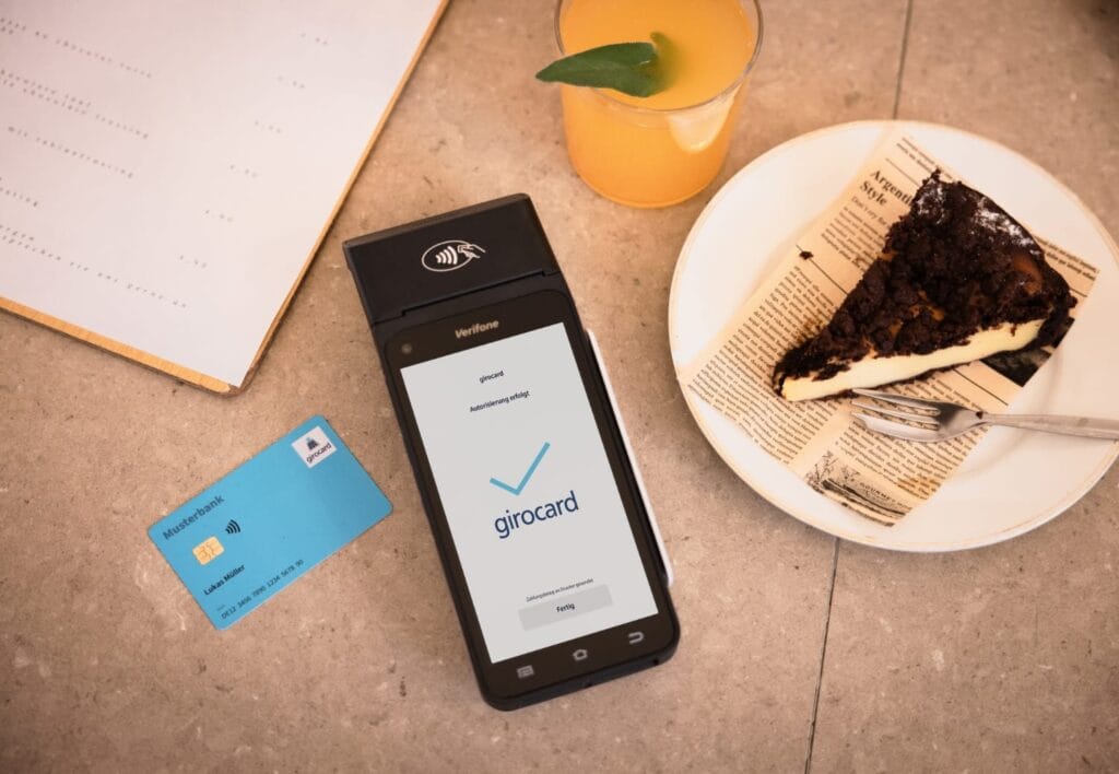 Verifone Payment-Terminal in Schwarz mit girocard NFC-Symbol, daneben eine blaue girocard und einbezahlsymbol, dazu ein Dessert und Getränk auf Tisch.