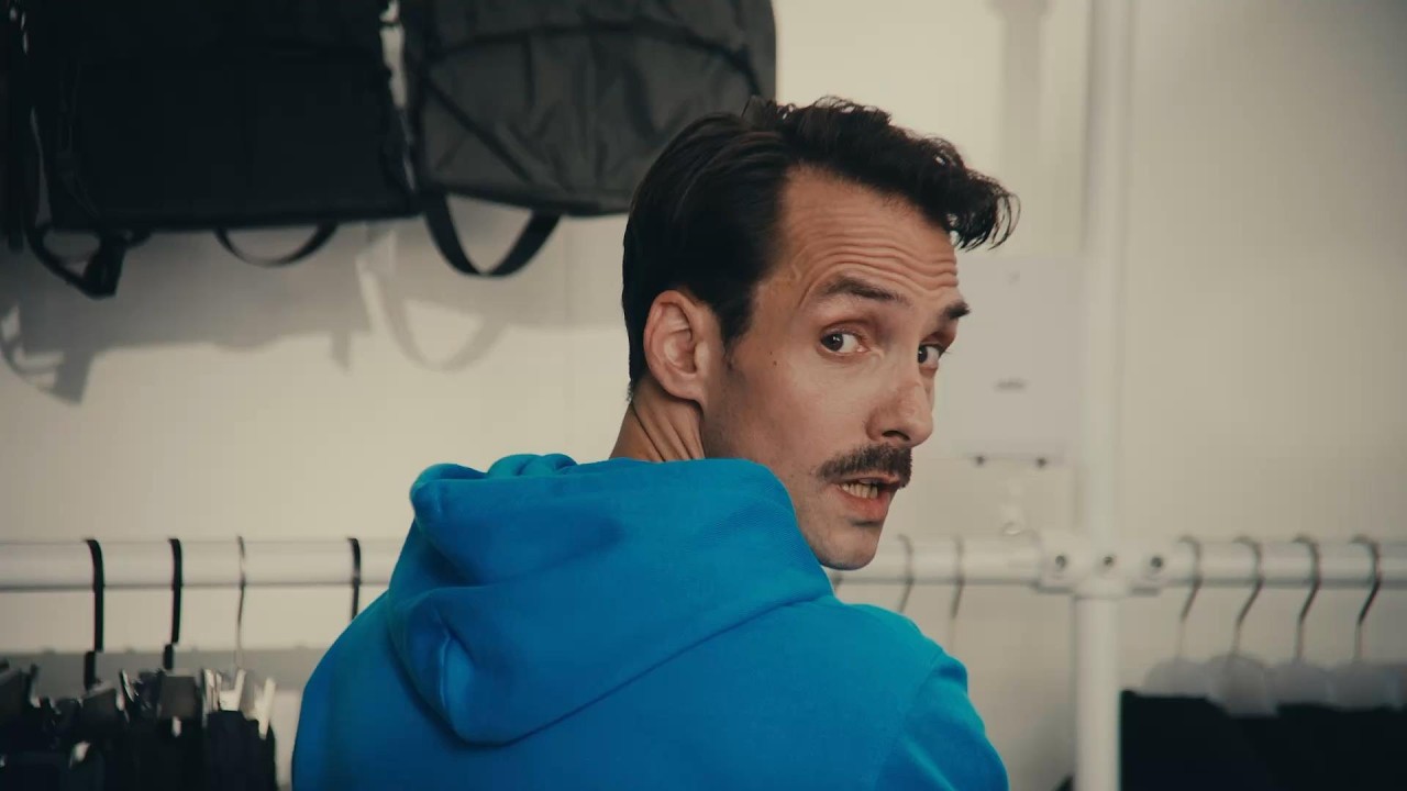 Mr. Giro dreht sich in einem Bekleidungsgeschäft zur Kamera um, im Hintergrund eine Kleiderstange.