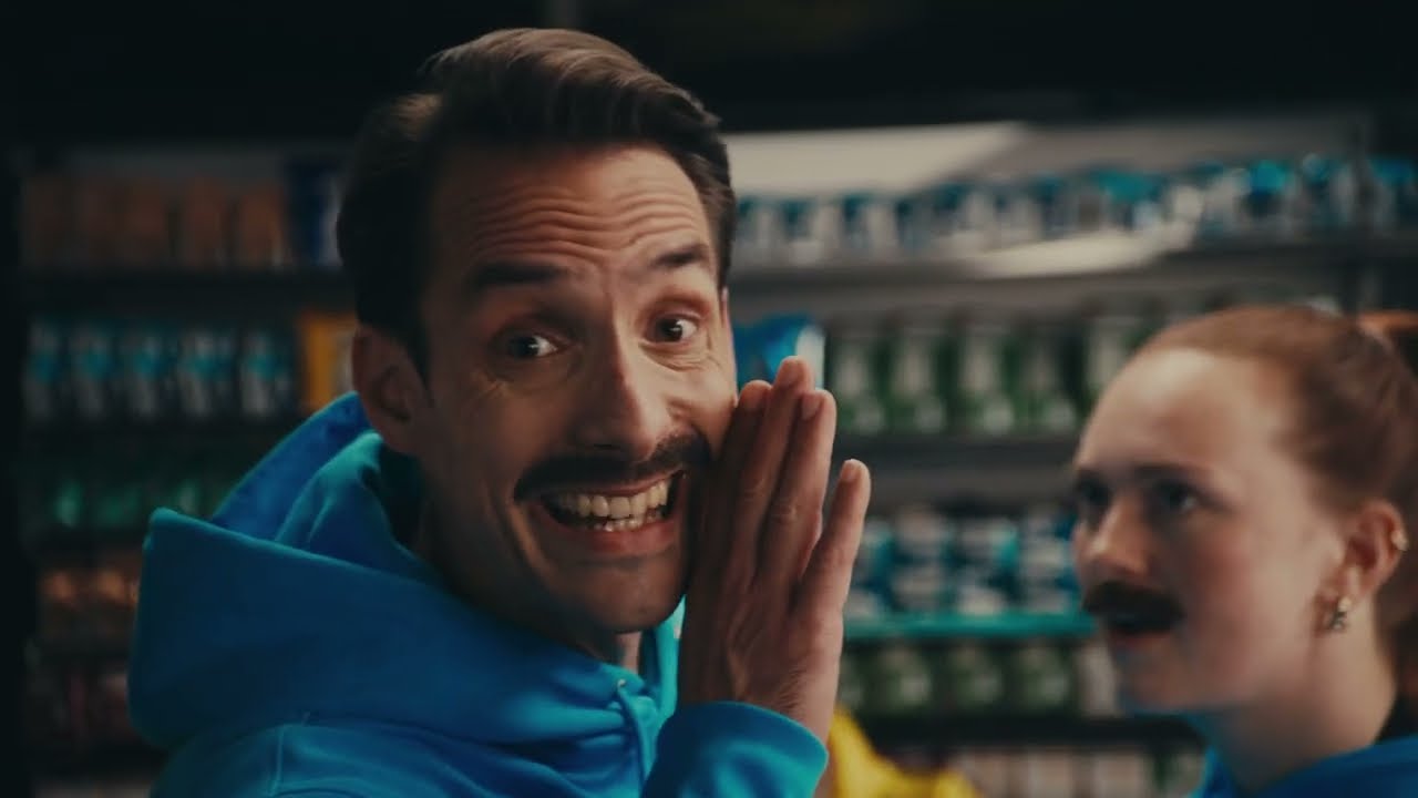 Mr. Giro hält die Hand an den Mund und flüstert, hinter ihm eine weitere Person mit Schnauzer im Späti.