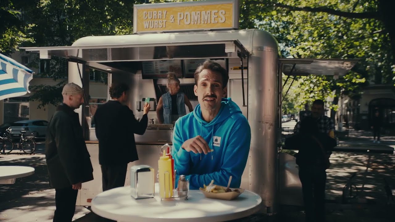 Mr. Giro sitzt an einem Stehtisch vor einer Currywurst-Bude, davor Senf und Ketchup.