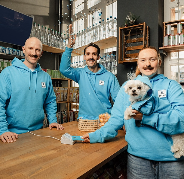Drei Mitarbeitende im girocard-Hoodie hinter der Ladentheke, eine Person hält einen kleinen Hund.